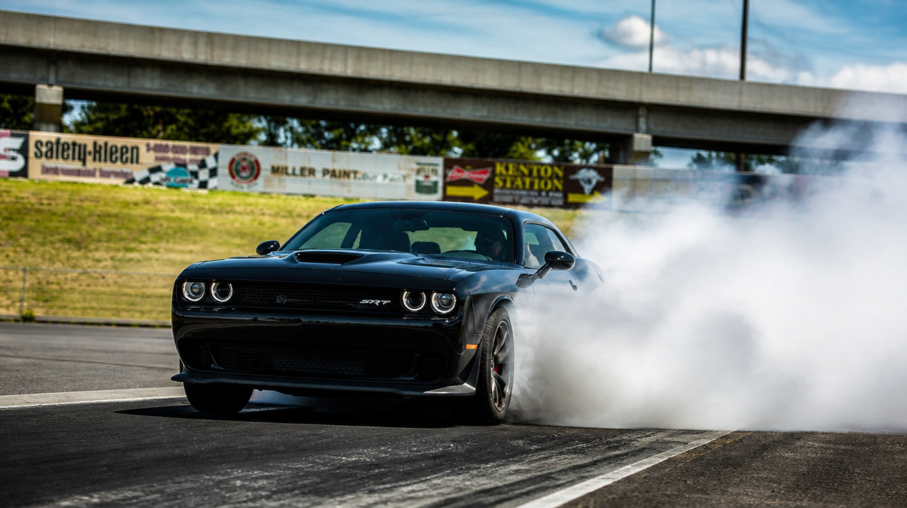 Schwarzer Dodge Challenger SRT driftet auf Rennstrecke, Reifen rauchen, Brücke und Werbetafeln im Hintergrund.