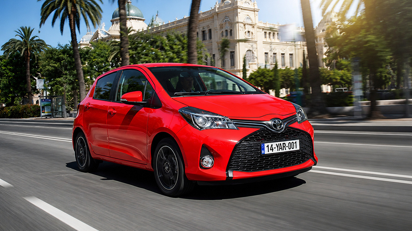 Roter Toyota Kleinwagen fährt auf sonniger Straße mit Palmen und historischem Gebäude im Hintergrund.