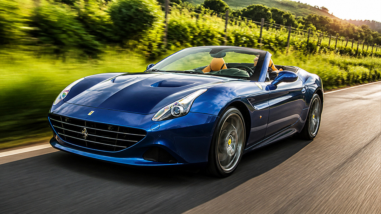 Blauer Ferrari California Cabriolet fährt auf Landstraße mit grüner Landschaft im Hintergrund.