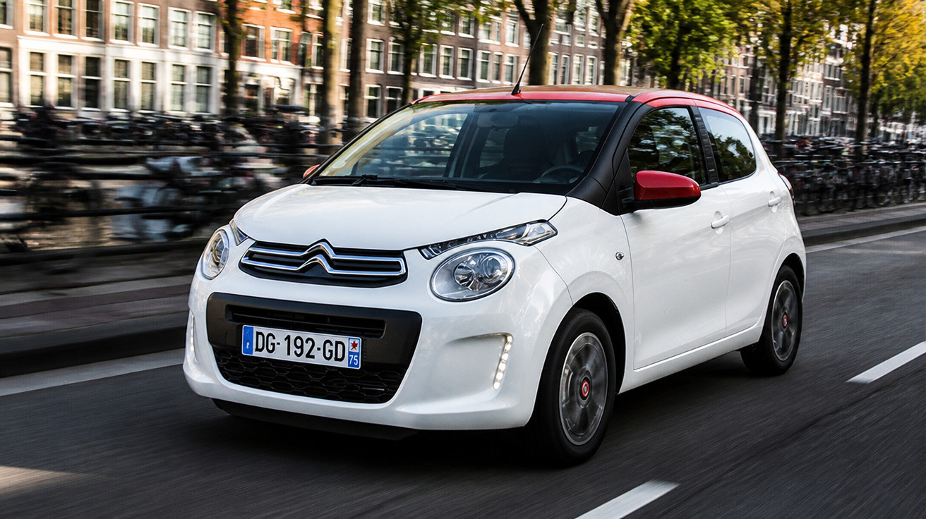Weißer Citroën Kleinwagen mit roten Außenspiegeln fährt auf Straße in städtischer Umgebung.