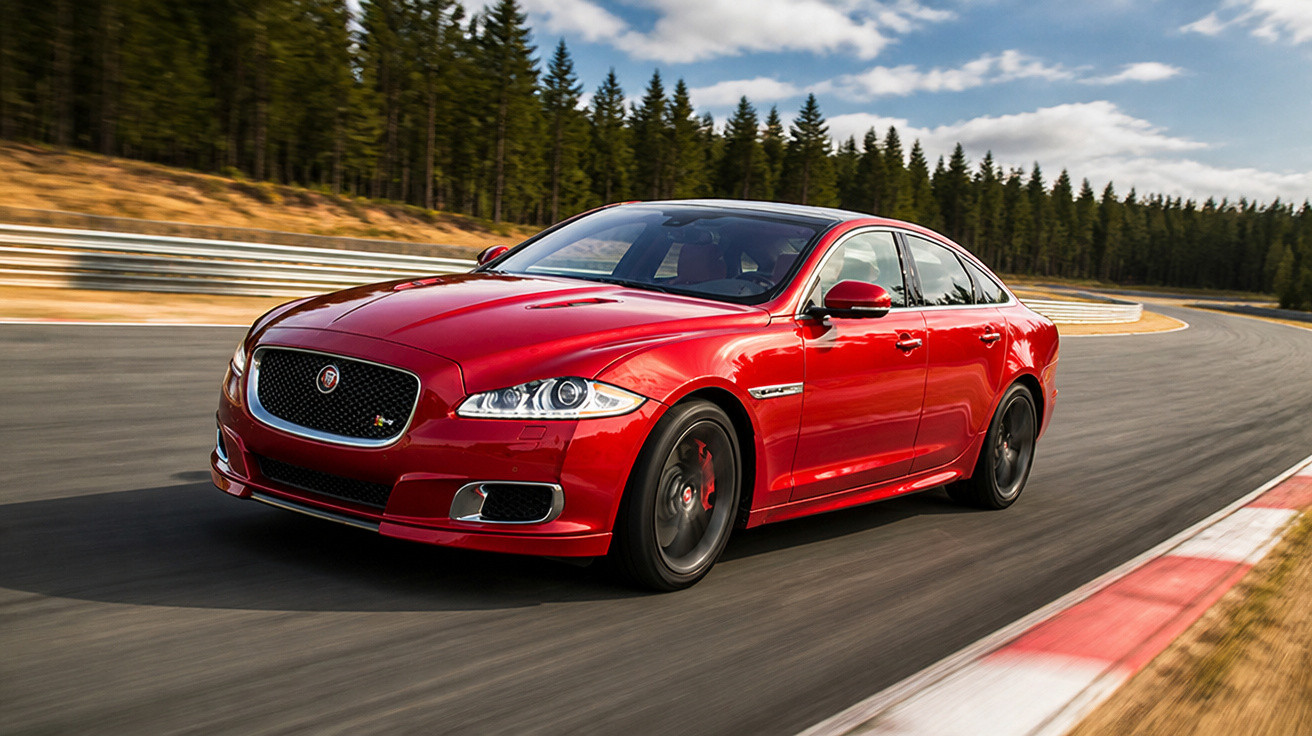 Roter Jaguar Sportwagen fährt auf Rennstrecke mit Wald im Hintergrund unter blauem Himmel.