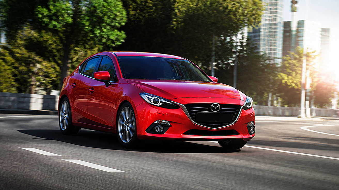 Roter Mazda fährt auf einer Straße mit Bäumen und Gebäuden im Hintergrund bei Sonnenschein.