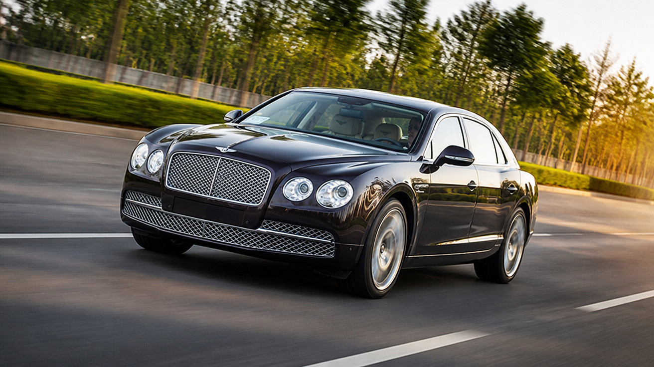 Schwarzer Bentley fährt auf einer Straße mit Bäumen im Hintergrund bei Sonnenlicht.