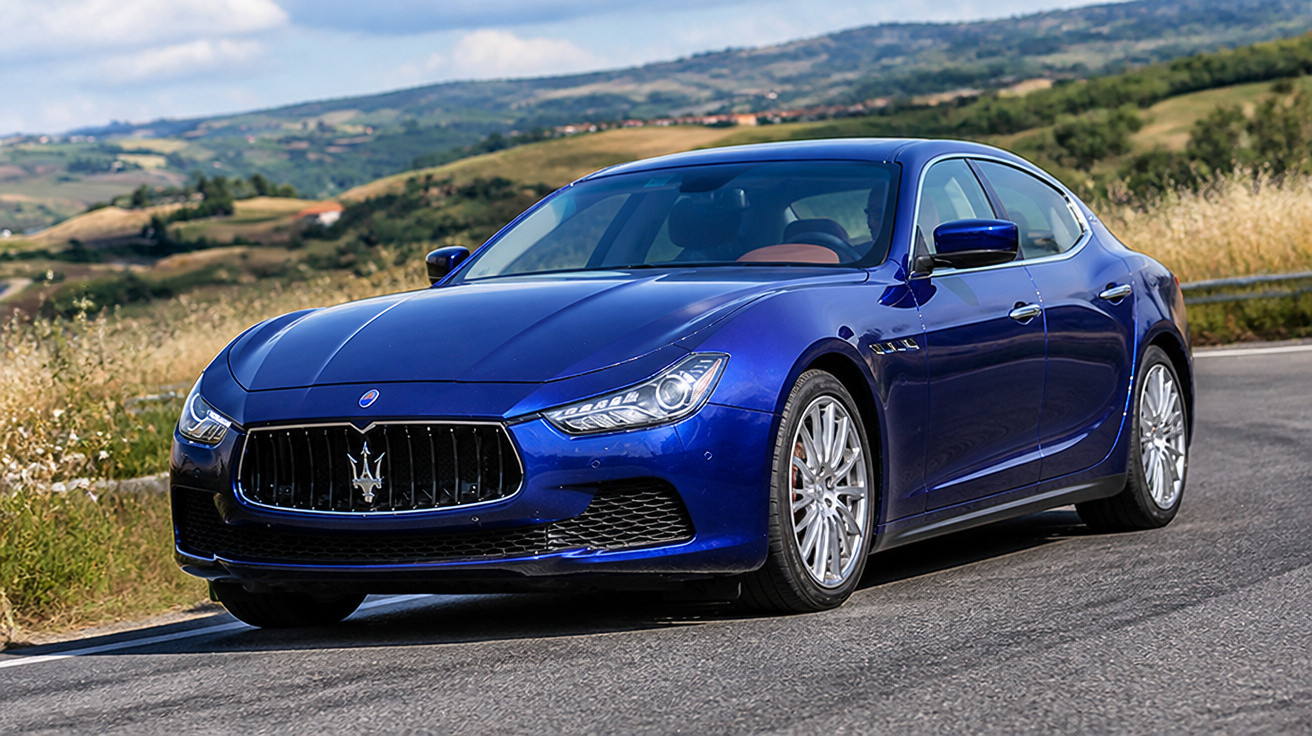 Blauer Maserati-Sportwagen auf Landstraße mit grünen Hügeln und bewölktem Himmel im Hintergrund.