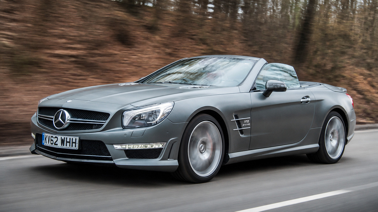 Silbergraues Mercedes-Benz Cabrio fährt auf einer Landstraße mit herbstlichem Waldrand im Hintergrund.